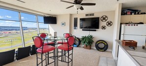 Una habitación con un ventanal grande, ventilador de techo, televisor, mesa con sillas y refrigerador.