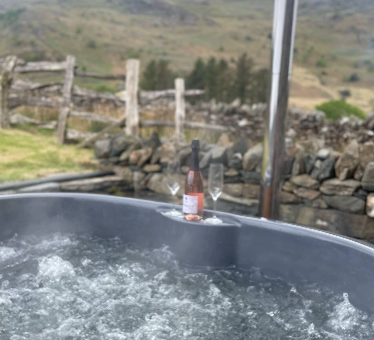 Ein Whirlpool mit einer Weinflasche und Gläsern, umgeben von einer Steinmauer und einem Holzzaun, vor einer Berglandschaft.