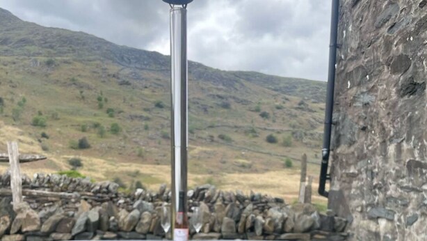 A wooden hot tub with a metal pole, set against a stone wall and a mountainous backdrop.
