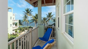 Ein Balkon mit zwei blauen Liegestühlen, einem weißen Geländer und Blick auf das Meer und Palmen.