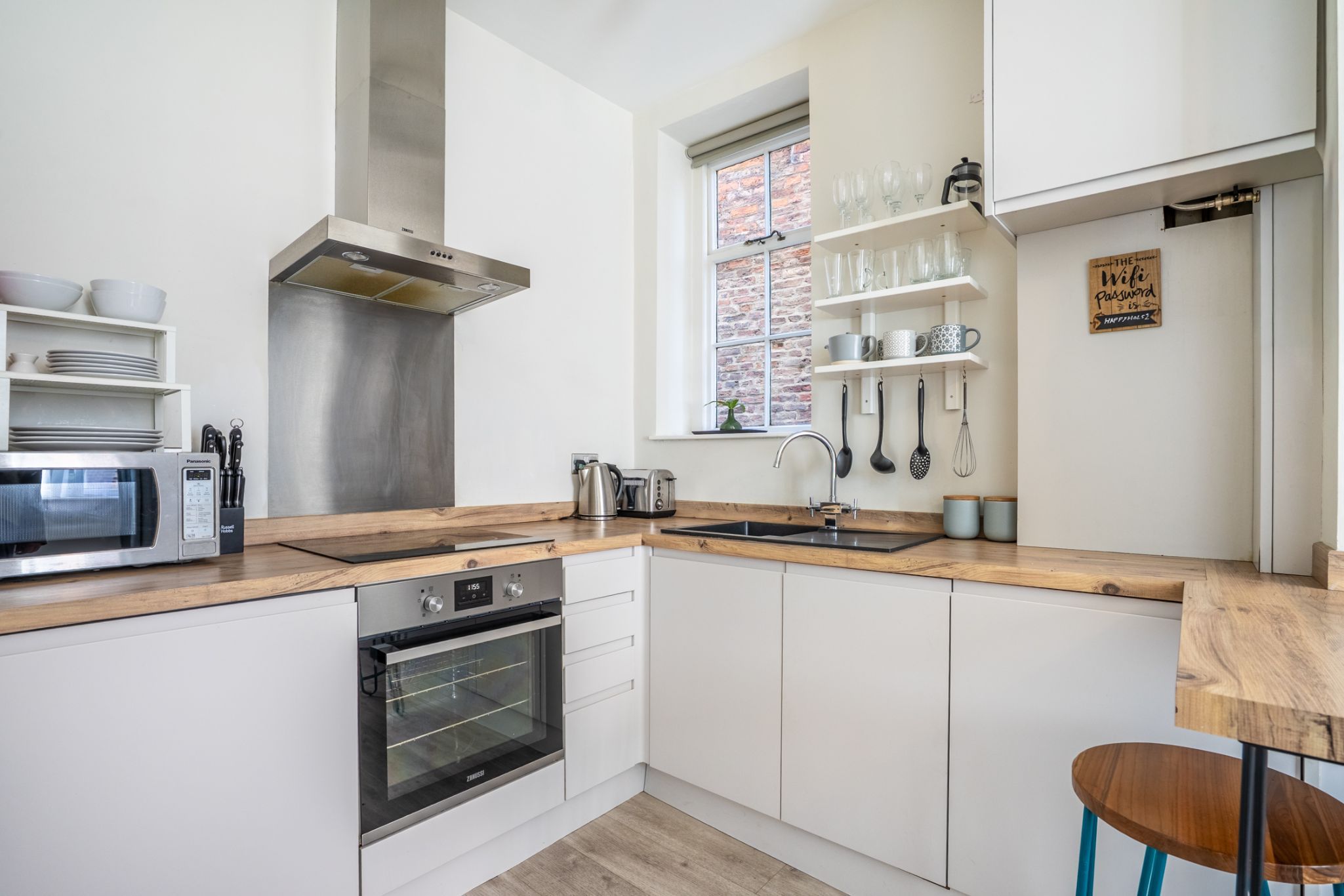 A modern kitchen with white cabinets, a stainless steel range hood, a built-in oven, a microwave, and a window with a brick pattern.