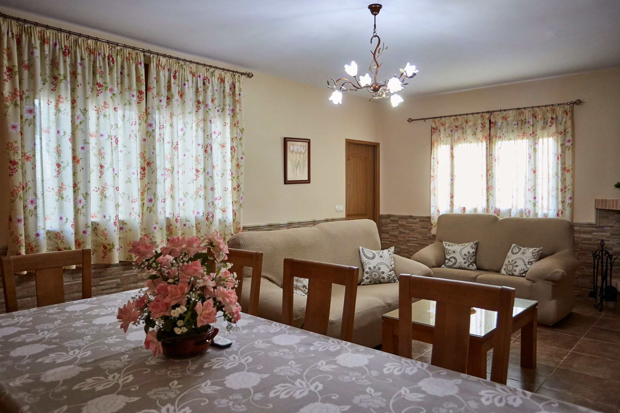 A living room with a beige sofa, wooden chairs, a dining table with a floral tablecloth, and a chandelier.