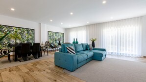 A modern living room with a sectional sofa, a dining table with chairs, and a large window with sheer curtains.