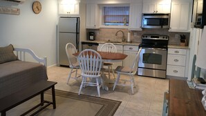 A compact kitchen with white cabinets, a stainless steel refrigerator, a microwave, and an oven. There is a dining table with four chairs and a bench. A clock is visible on the wall. - Cozy Studio in Long Beach / EAB (East Atlantic Beach)