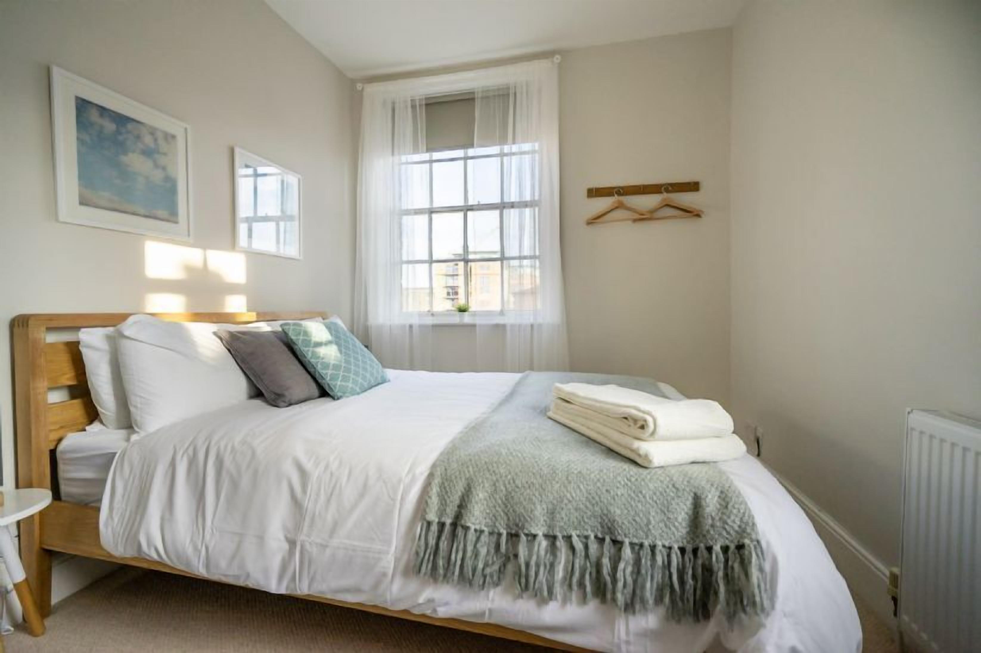 A neatly made bed with white linens and a grey throw blanket, a bedside table with a lamp, and a window with sheer curtains.