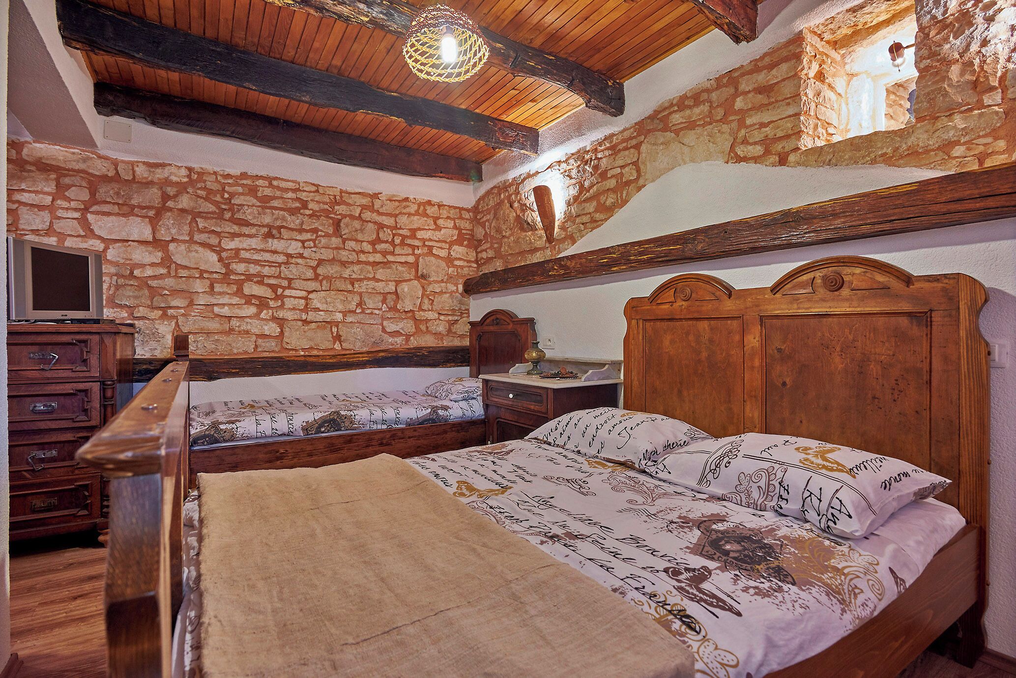 Ein Schlafzimmer mit Stehwänden, Holzbalken, einem Bett mit gemusterten Betttextilien, einer hölzernen Kommode und einem kleinen Fenster.