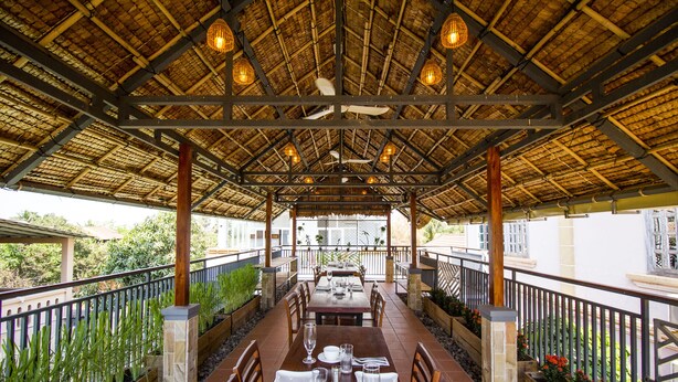 An outdoor dining area with tables set for four, under a thatched roof with hanging lights. - Koki Tree (Siem Reap)