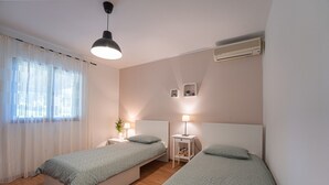 Un dormitorio con dos camas individuales, una mesita de noche con lámpara, un pequeño estante con plantas y un equipo de aire acondicionado en la pared.