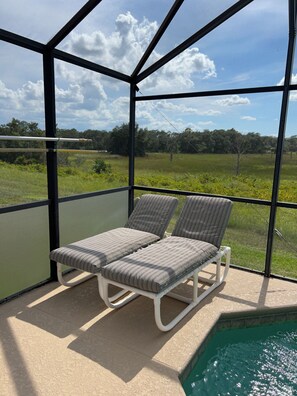 A balcony with two striped lounge chairs, a pool, and a view of a grassy field and trees.