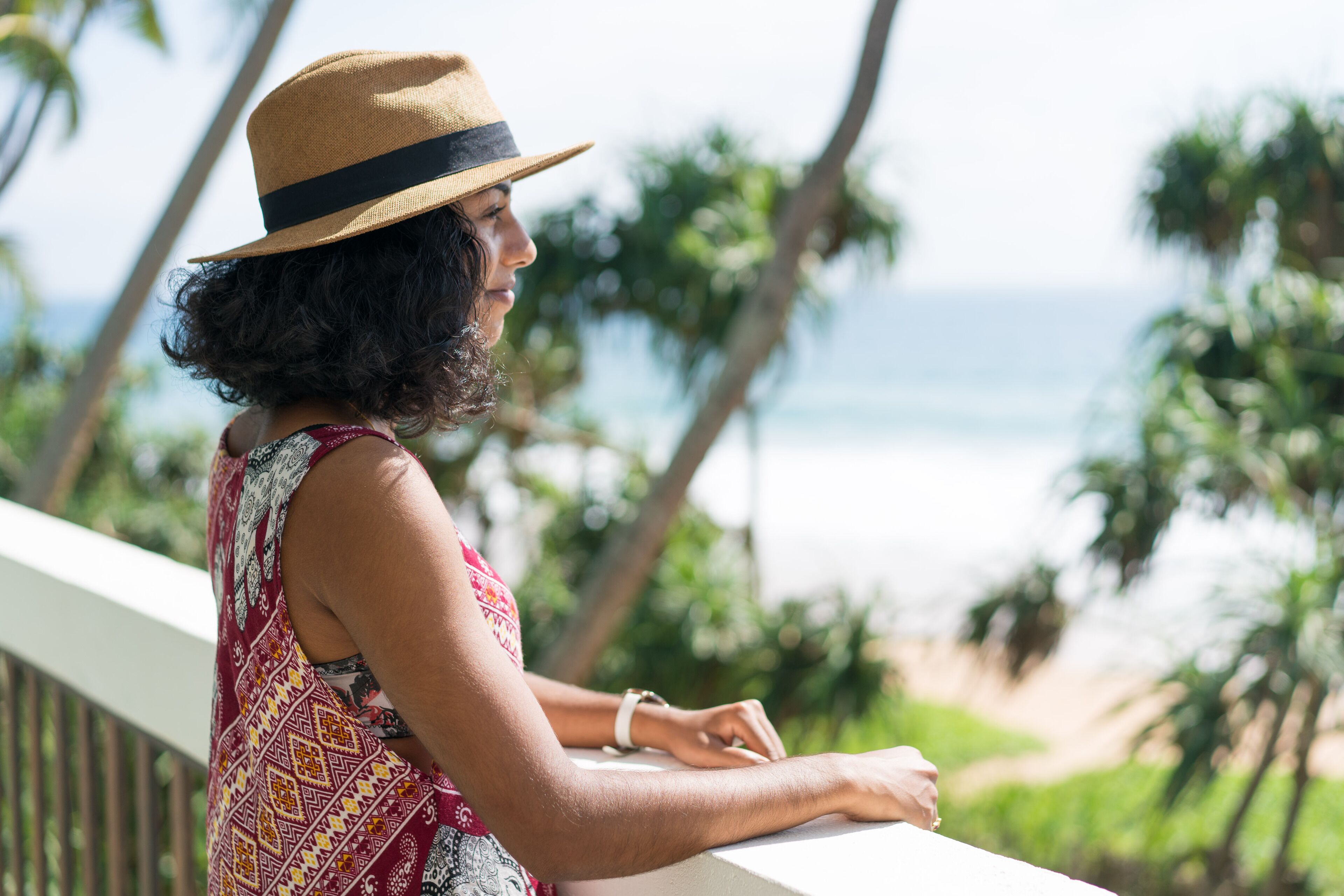 A person leaning on a balcony railing overlooking a beach with palm trees and the ocean.