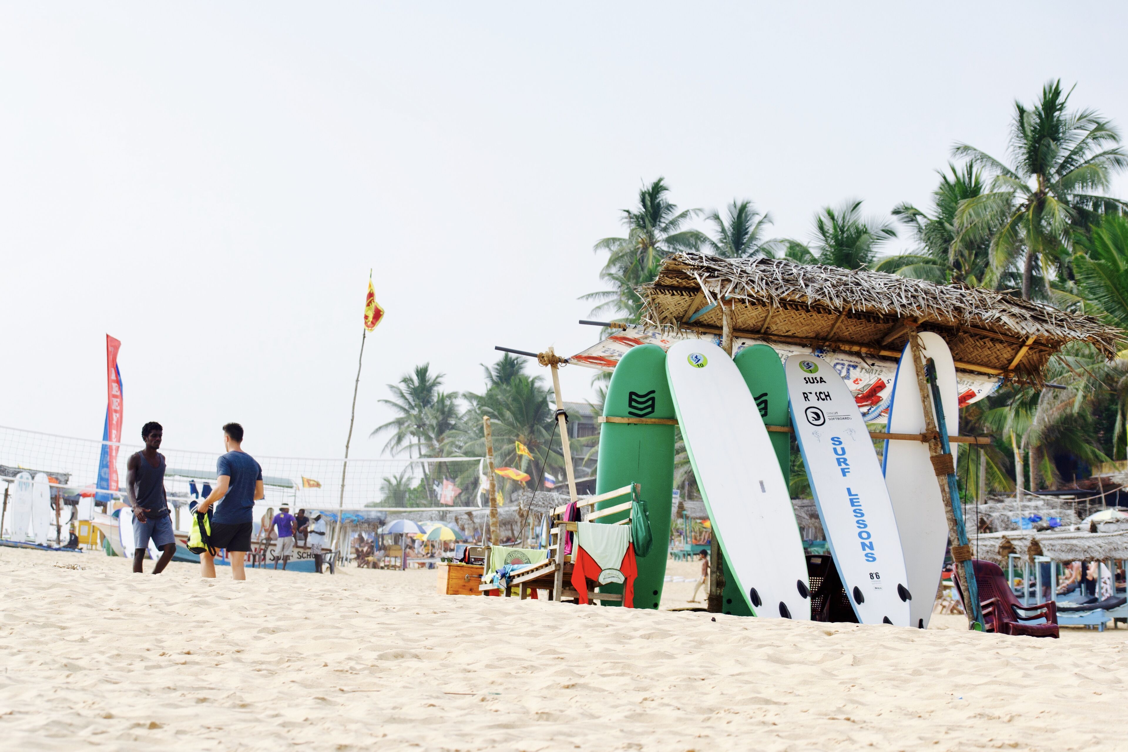 A beach with surfboards leaning against a hut, palm trees, and beach umbrellas.