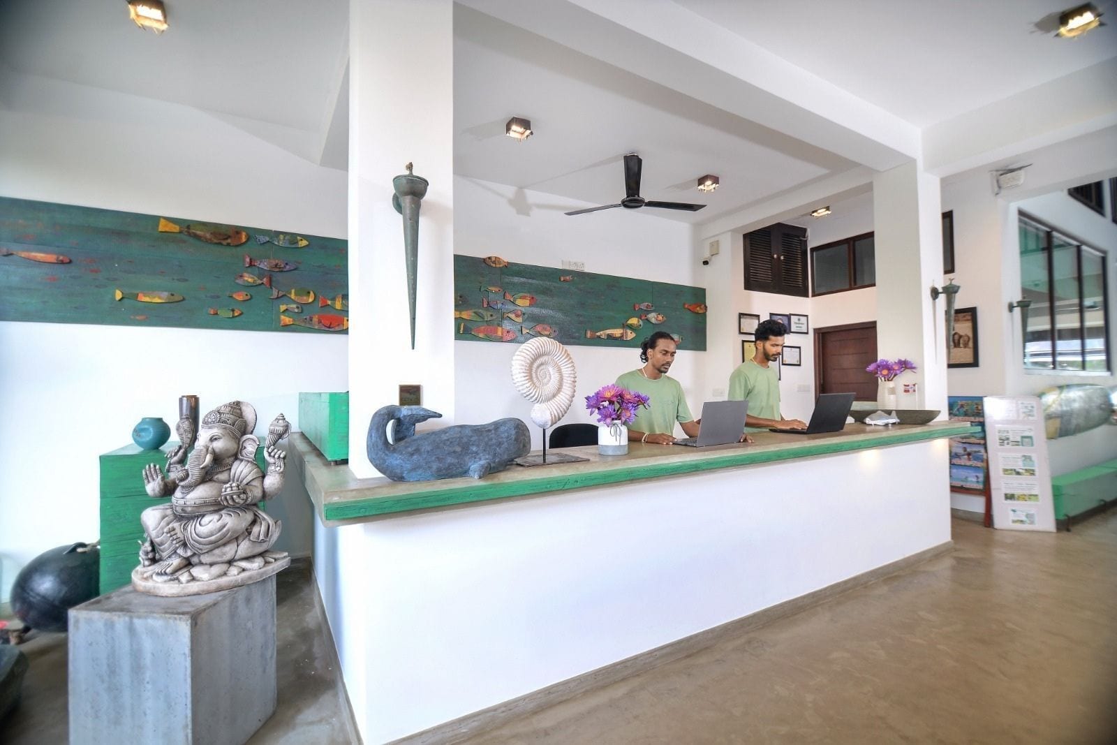 A reception desk with two staff members, a ceiling fan, artwork on the walls, and a green accent wall.