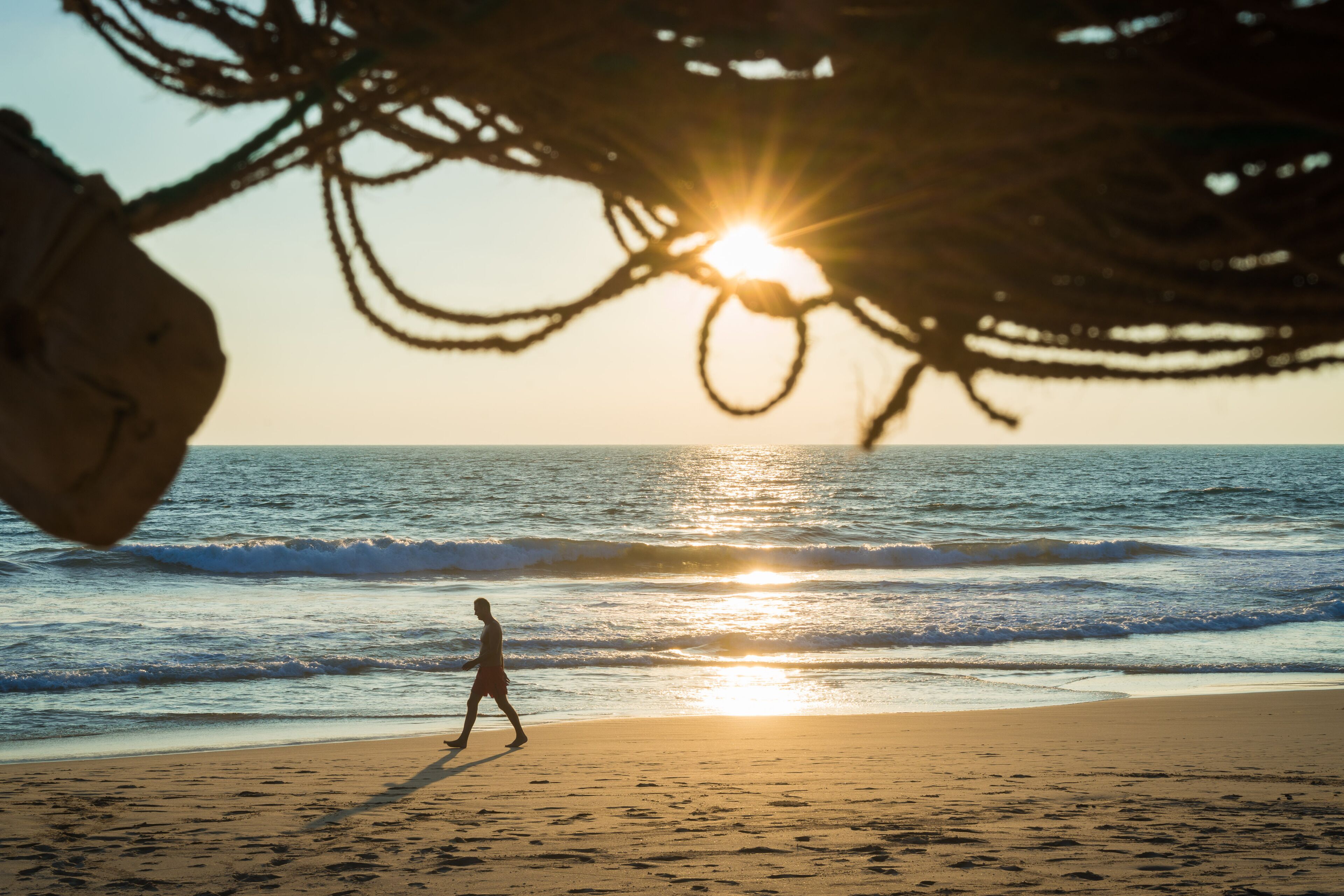 Une scène de plage où l’on voit une personne marchant en direction du coucher de soleil, avec des vagues en arrière-plan et une structure faite de cordes au-dessus.