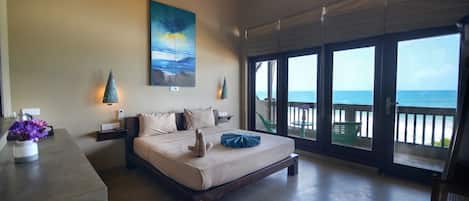 A bedroom with a bed, a nightstand, a vase with flowers, a painting, and a balcony with a view of the ocean.