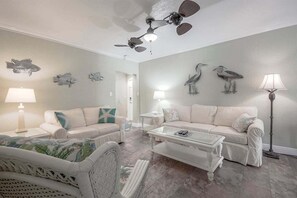 A living room with a white sofa, a coffee table, and a ceiling fan. - Stunning Beach Suite, Heated Pool, Steps to Sand! Clearwater ➠ Beach Suites #105 (Clearwater Beach)