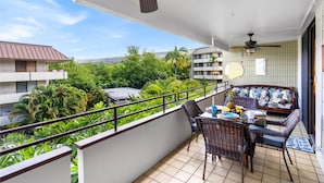 A balcony with wicker furniture, a ceiling fan, and a view of a lush green landscape. - 🏖️Across from Magic Sands Beach | Kona 2BR Condo w/ AC, Lanai, Pool & Sauna (Kailua Kona)