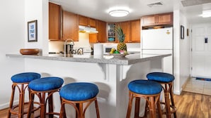 A kitchen with wooden cabinets, a white refrigerator, and a central island with stools. - 🏖️Across from Magic Sands Beach | Kona 2BR Condo w/ AC, Lanai, Pool & Sauna (Kailua Kona)