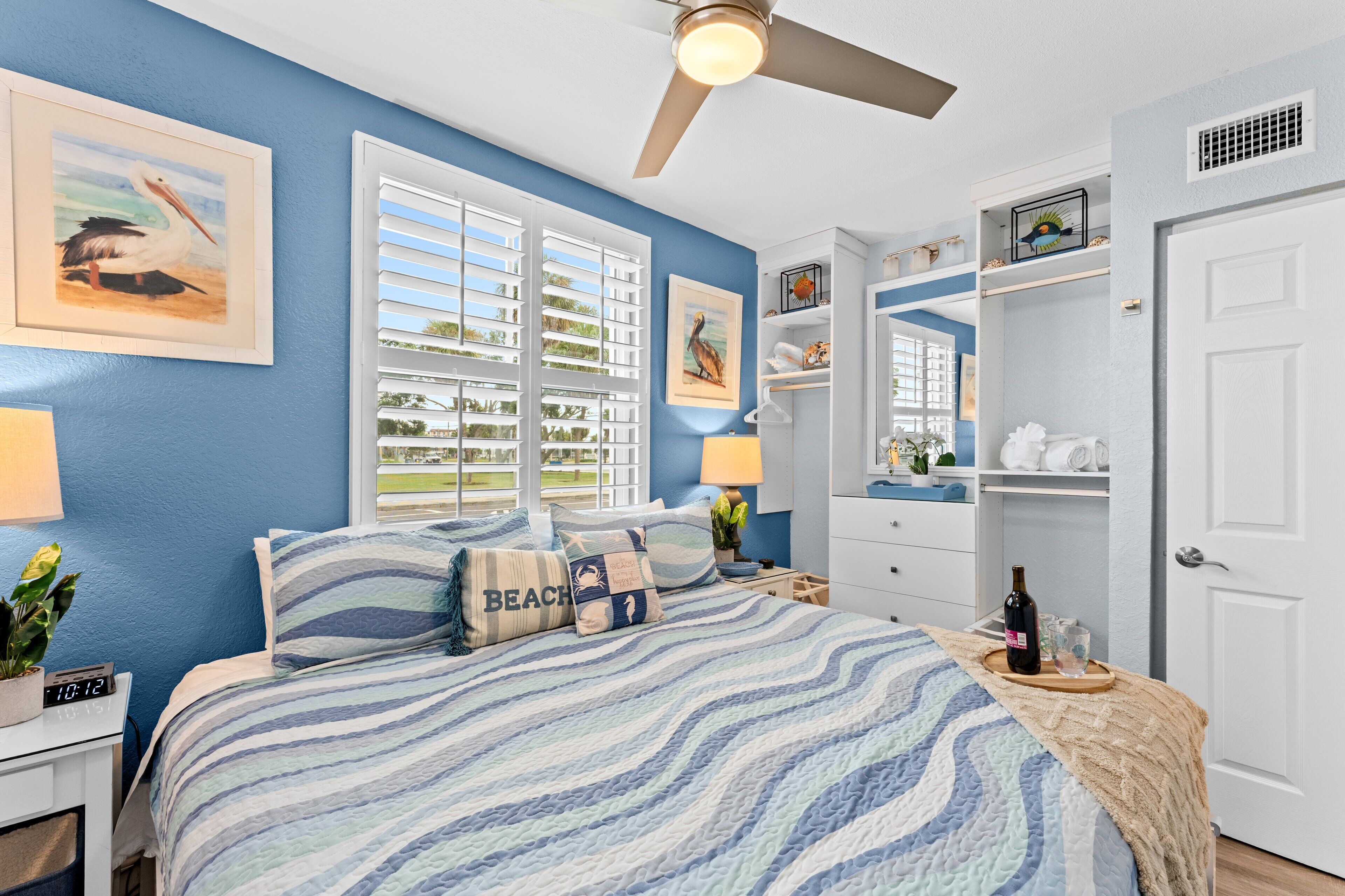 Une chambre à coucher comprenant un lit, une table de chevet, une lampe, un ventilateur de plafond et une fenêtre avec des stores.