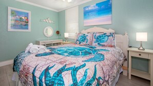 A bedroom with a bed, a ceiling fan, a nightstand, a lamp, and artwork on the walls. - Cute beach condo with heated pool, steps to the sand ➠ CBS 204 (Clearwater)