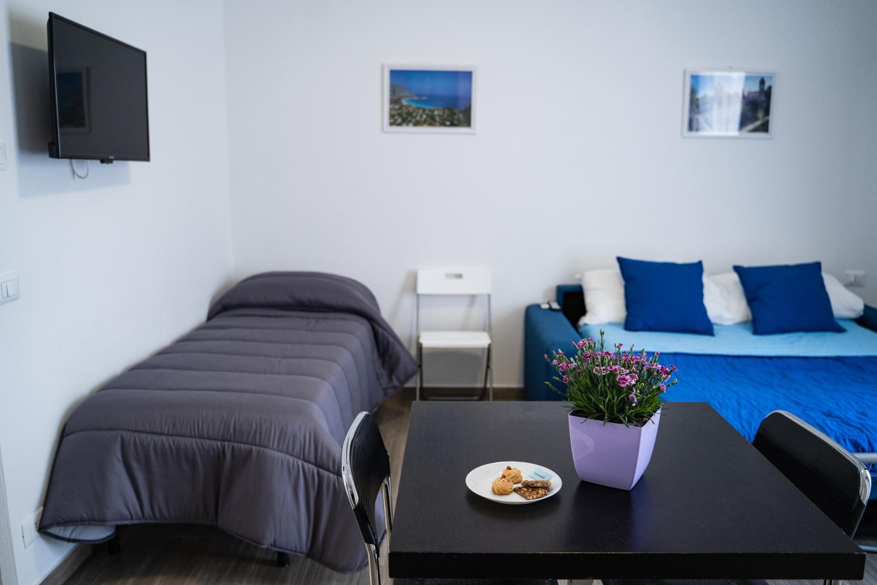 Un dormitorio con cama, una mesita con un plato de comida y una maceta, un televisor montado en la pared y dos cuadros enmarcados.