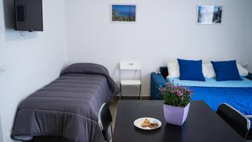 Un dormitorio con cama, una mesita con un plato de comida y una maceta, un televisor montado en la pared y dos cuadros enmarcados.