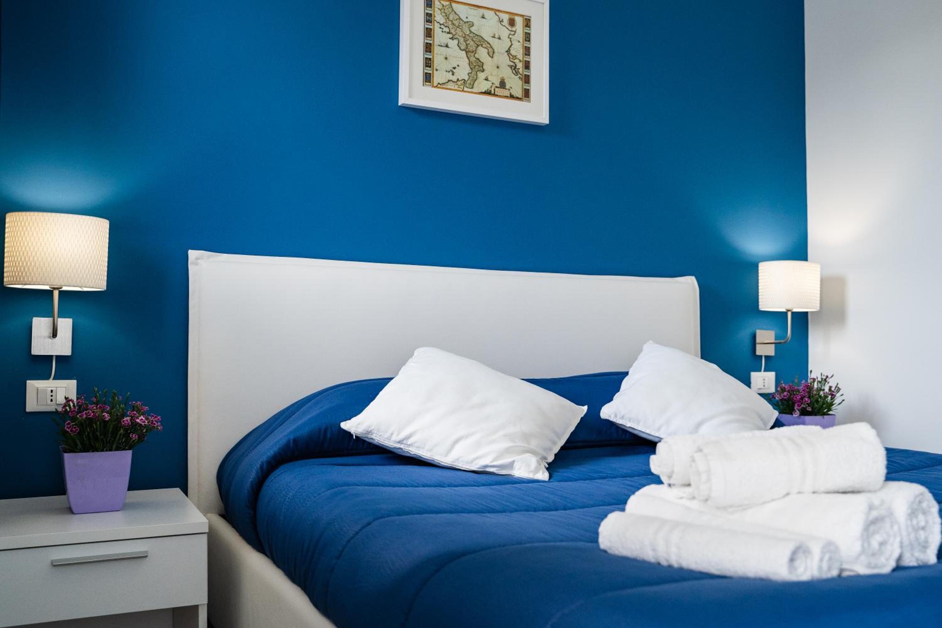 A neatly made bed with white pillows and rolled towels, a blue wall, and a framed map.