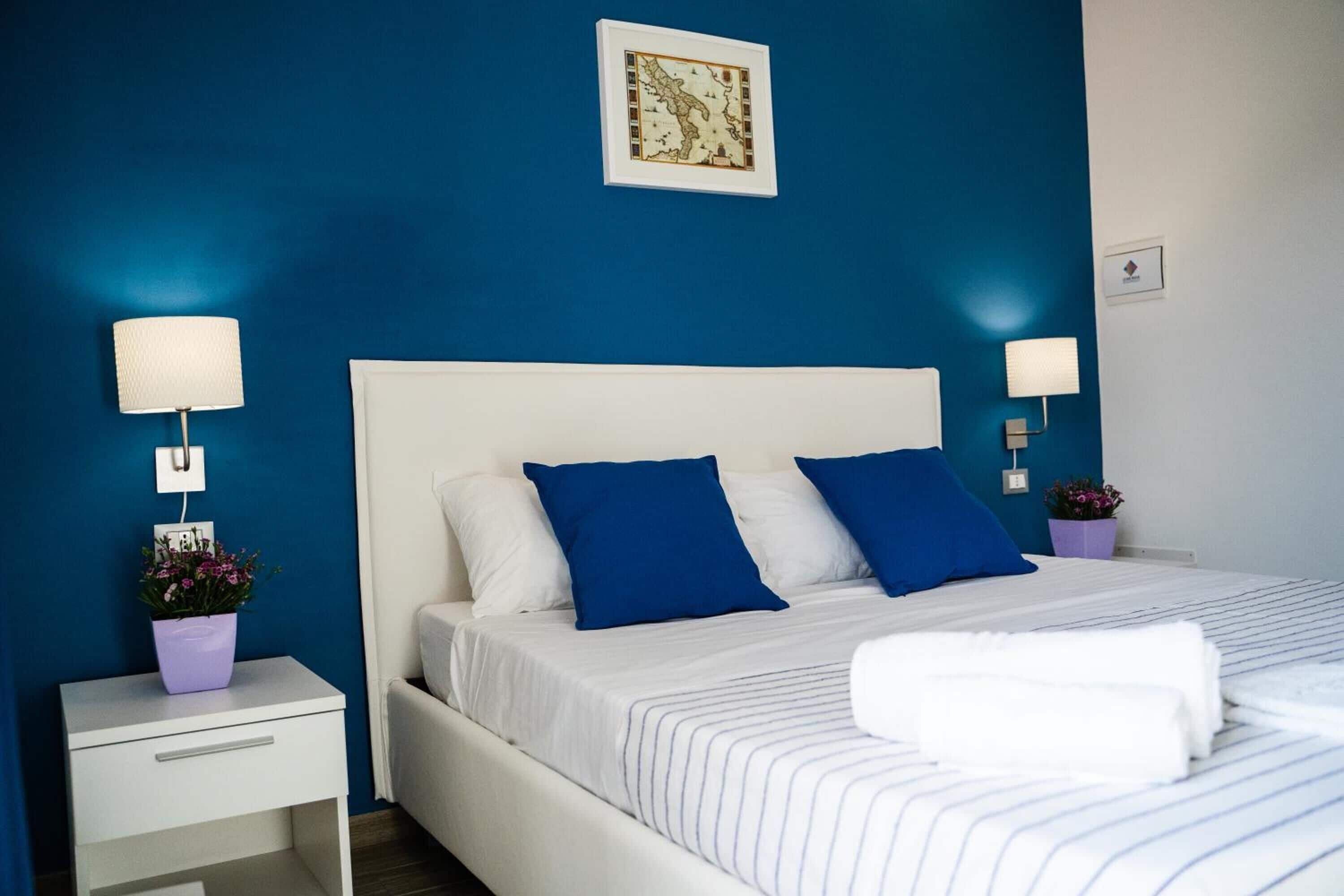 Una cama bien hecha con ropa de cama azul y blanca, dos lámparas de noche y una pequeña planta en maceta sobre la mesita.