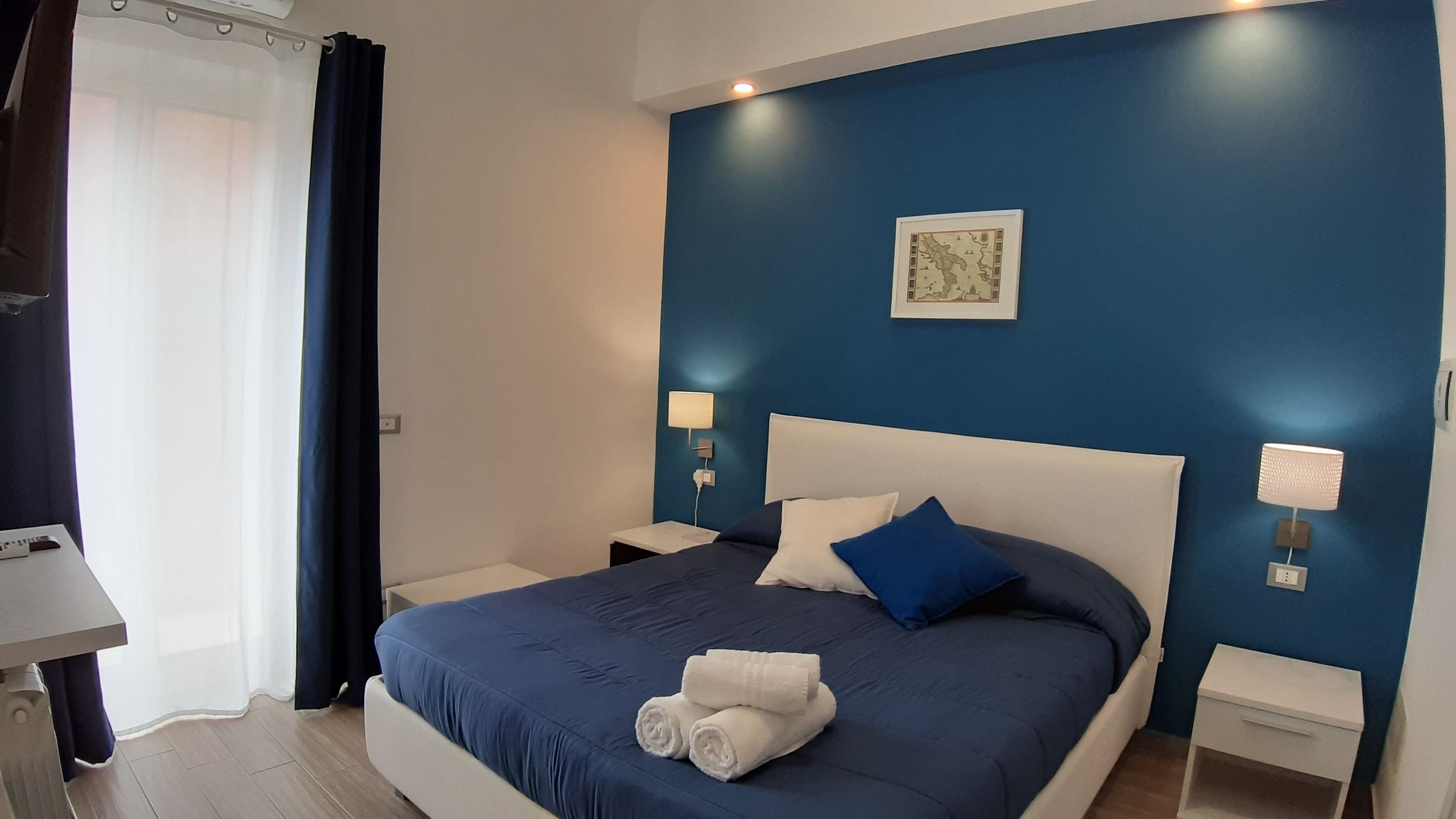 Ein ordentlich bezogenes Bett mit blauer Bettdecke, weißen Kissen und einem blauen Zierkissen. Zwei Nachttische mit Lampen, eine gerahmte Karte an der Wand und ein Paar zusammengerollte Handtücher auf dem Bett.