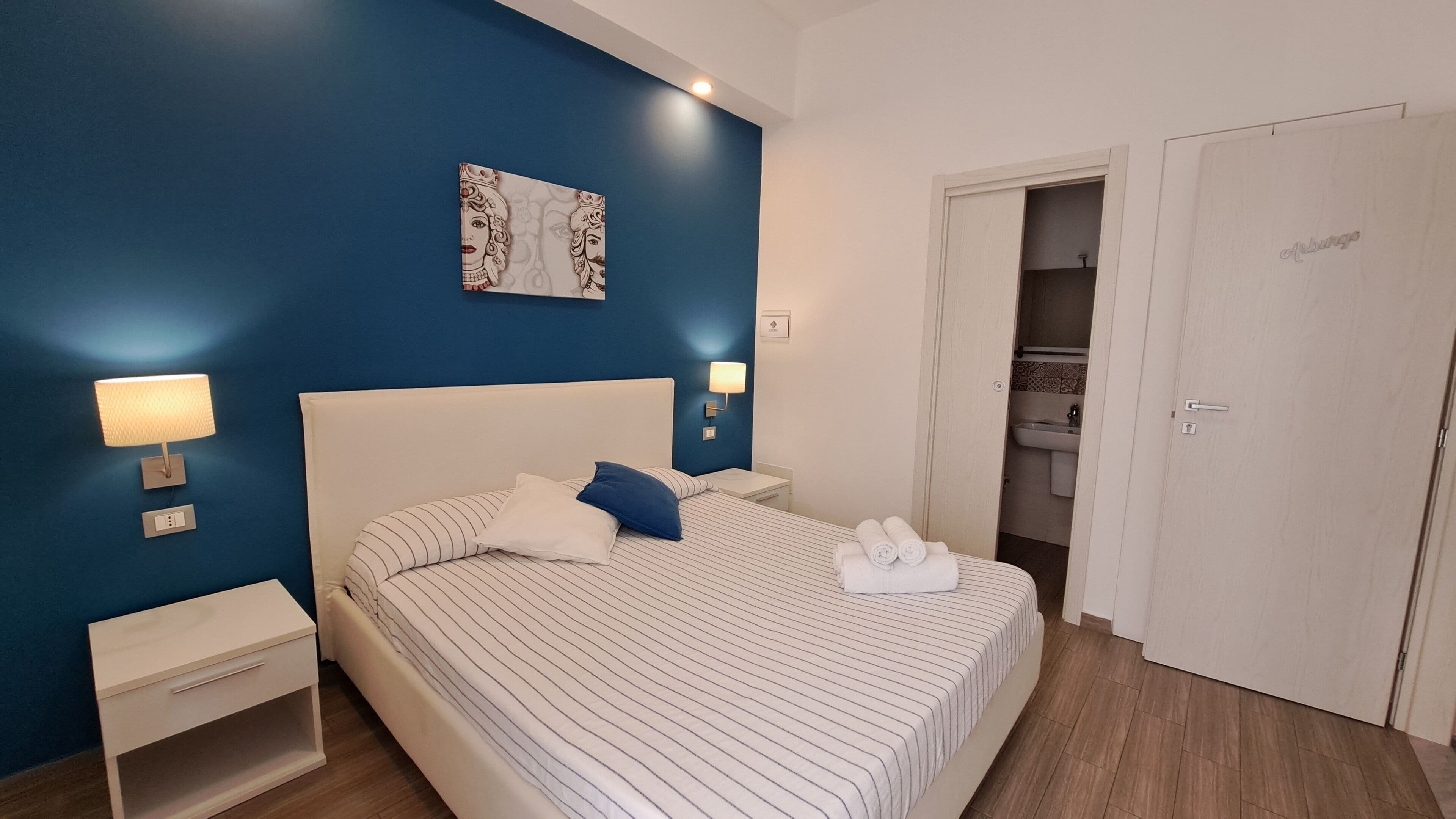 A hotel room with a large bed, two bedside tables, a wall-mounted lamp, and a door leading to a bathroom.