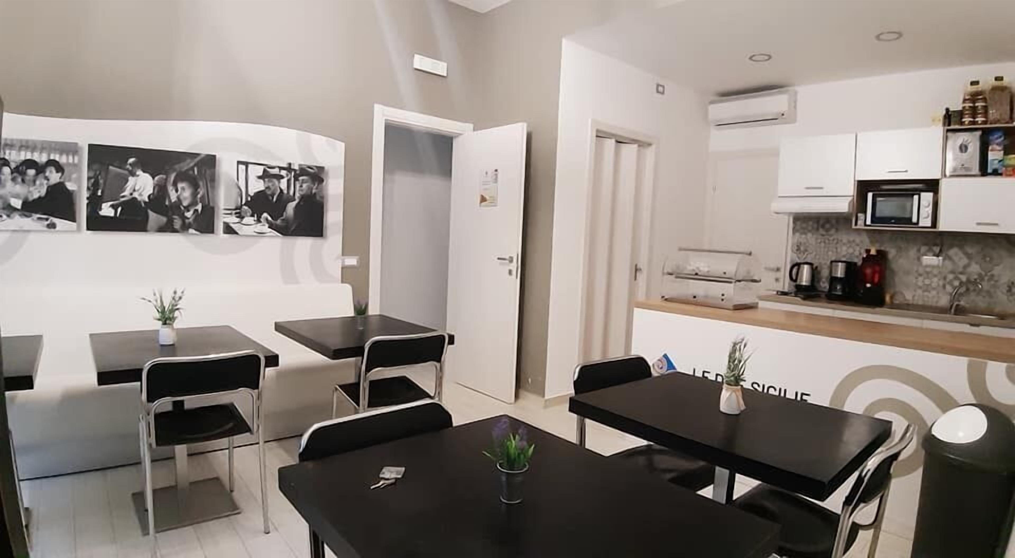 Una cocina moderna con armarios blancos, un microondas y una cafetera. Hay dos mesas negras con sillas, y una puerta que da a otra habitación.