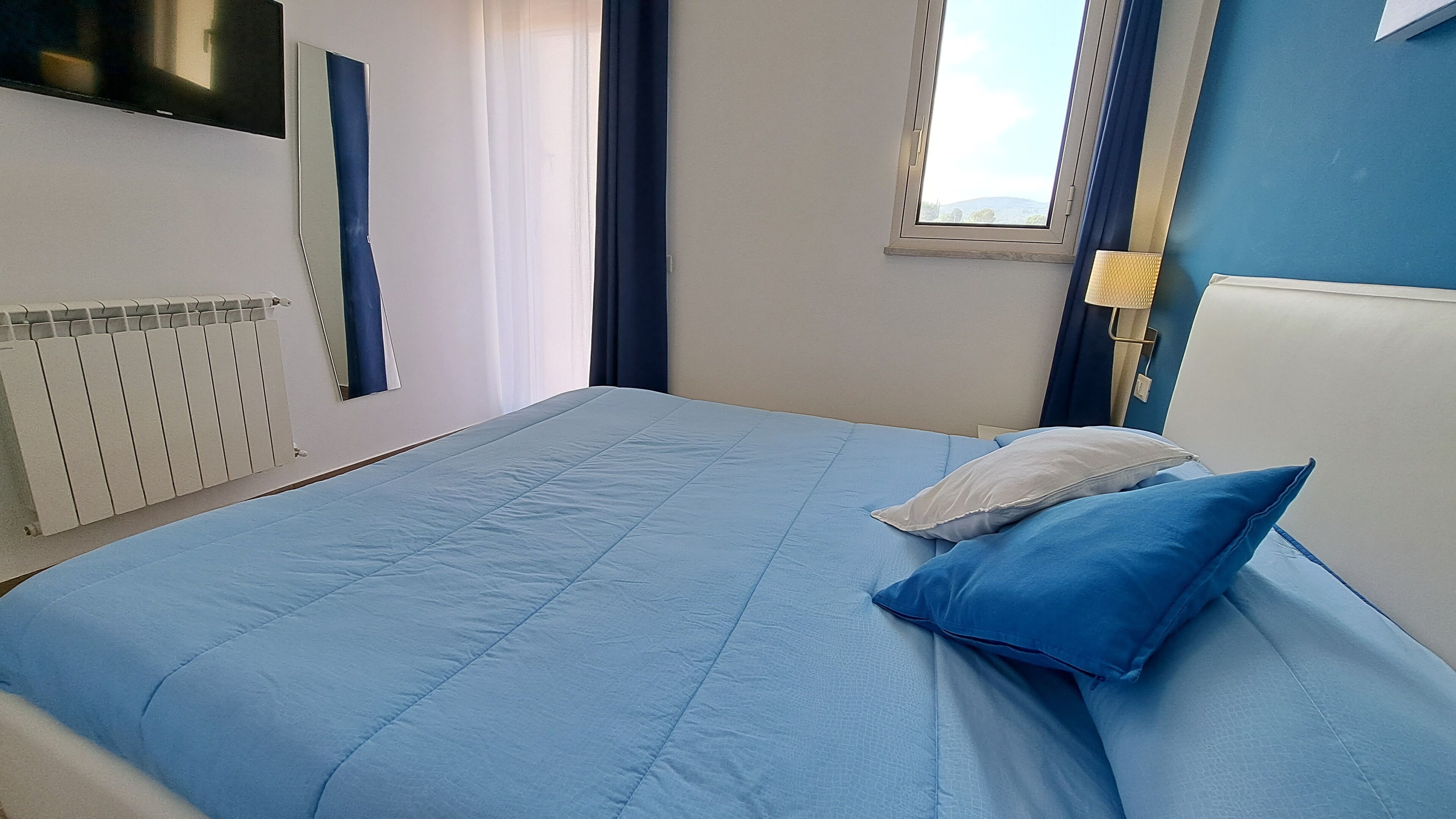 A hotel room with a large bed, a window with a view, a radiator, and a lamp.