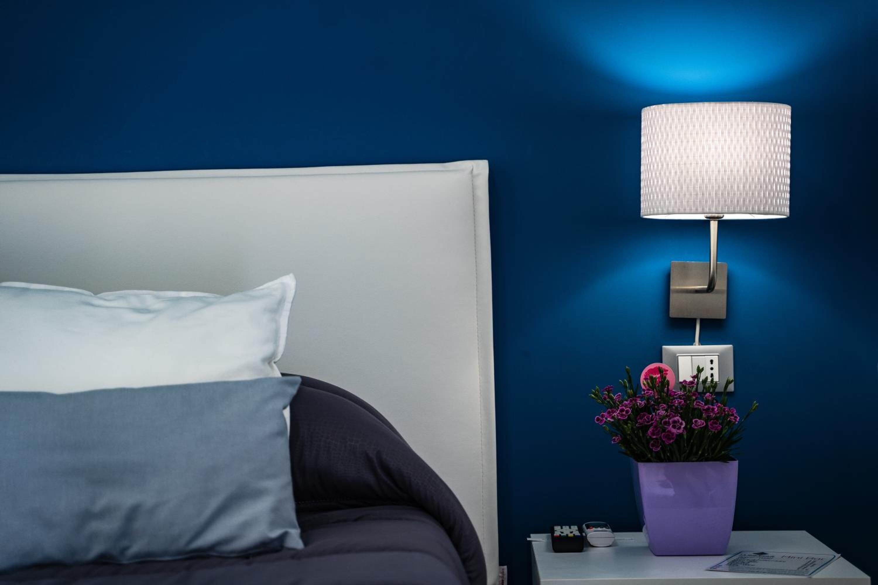 Ein ordentlich bezogenes Bett mit weißem Kopfteil, ein Nachttisch mit Lampe, ein violettes Blumentöpfchen und eine Steckdose an der Wand.
