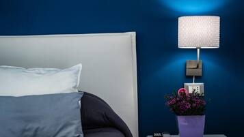 Ein ordentlich bezogenes Bett mit weißem Kopfteil, ein Nachttisch mit Lampe, ein violettes Blumentöpfchen und eine Steckdose an der Wand.