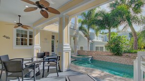 Un espace aménagé au bord de la piscine, avec une table et des chaises, un ventilateur de plafond, et une vue sur la piscine et les palmiers.