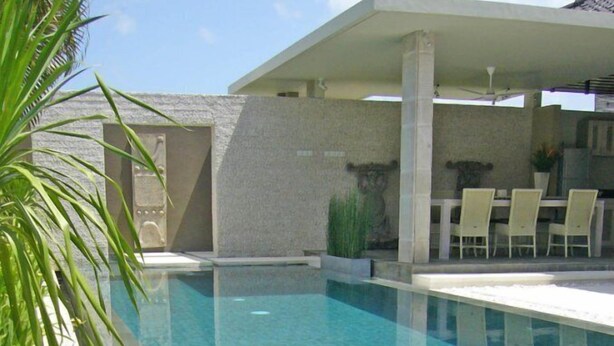 A swimming pool with clear water, surrounded by a concrete deck and a covered seating area with chairs.