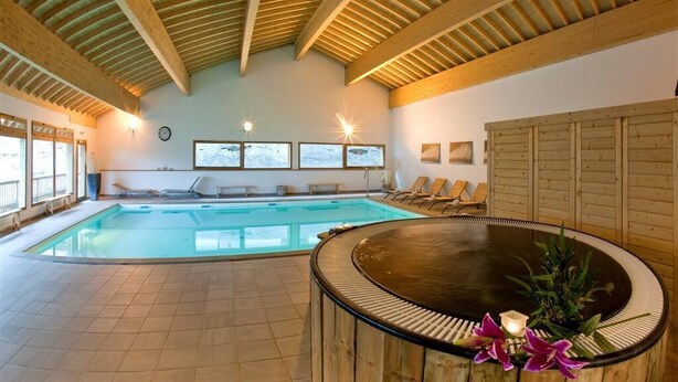 Una piscina interior con techo de madera, un jacuzzi y ventanas iluminadas por el sol.