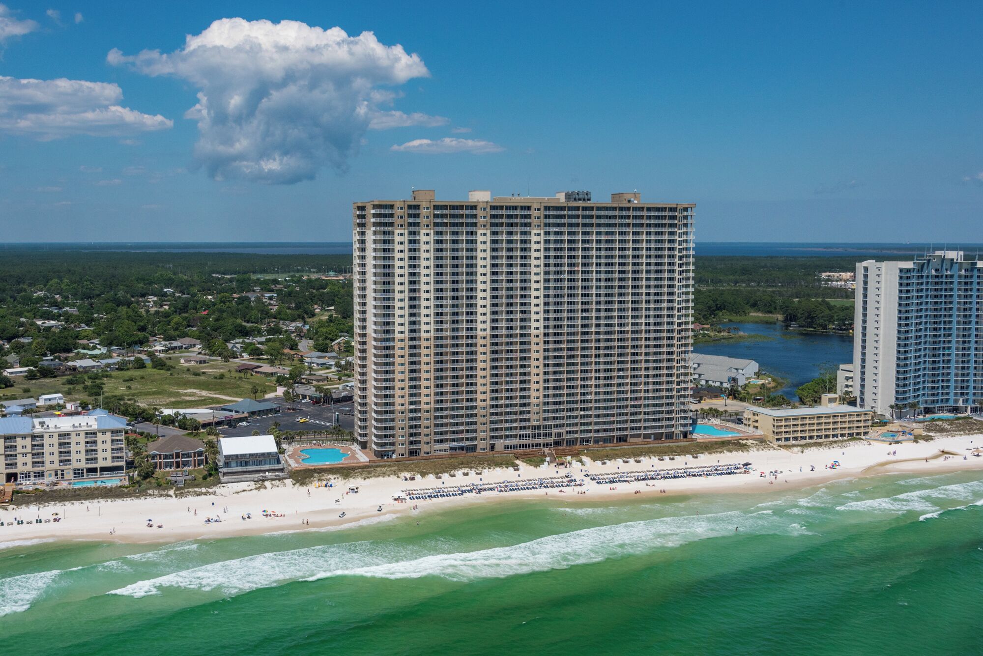A high-rise hotel with multiple floors, a beach in the foreground, and a body of water in the background.
