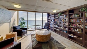 Una biblioteca amb un gran finestral que dona al mar, amb prestatgeries, una cadira de pell i un otomà de vímet.