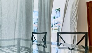 A glass-top table with a geometric metal frame, a white curtain, and a view of buildings and palm trees outside. - Los Corales Beach Village - Your private paradise - 2BR first floor - LT#D2b2 (Bávaro)