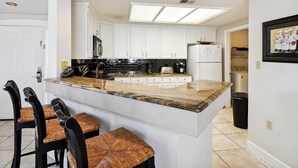 Uma cozinha moderna com armários brancos, uma ilha central com banquetas e uma janela grande acima da pia.