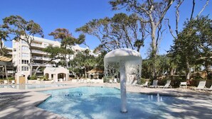 Piscina com uma fonte, espreguiçadeiras e um edifício com varandas ao fundo.