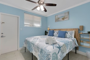 A bedroom with a bed, a ceiling fan, a window with blinds, a wall-mounted shelf, and a door.