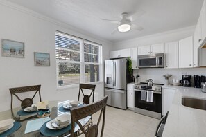 A modern kitchen with white cabinets, stainless steel appliances, a dining table with chairs, and a large window with blinds.