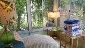 A cozy room with a bed, a lamp, a wicker chair, and a table with folded blankets. - "OLD FLORIDA" RIVERSIDE POOL COTTAGE THAT TIME FORGOT (Sebastian)