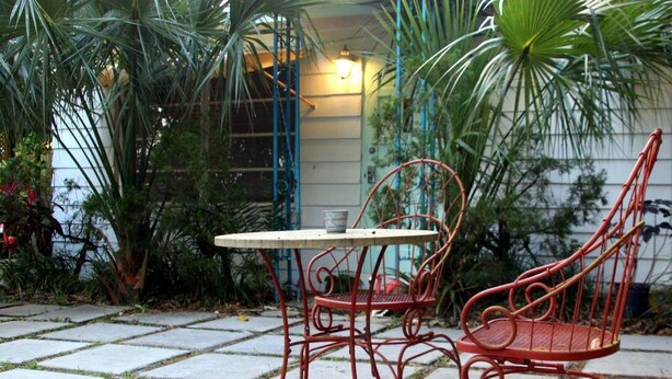 A patio with a metal table and two chairs, surrounded by greenery and a building in the background. - "OLD FLORIDA" RIVERSIDE POOL COTTAGE THAT TIME FORGOT (Sebastian)