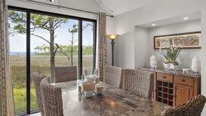 Een eetgedeelte met een grote tafel, rieten stoelen en uitzicht op een grasveld door grote ramen.