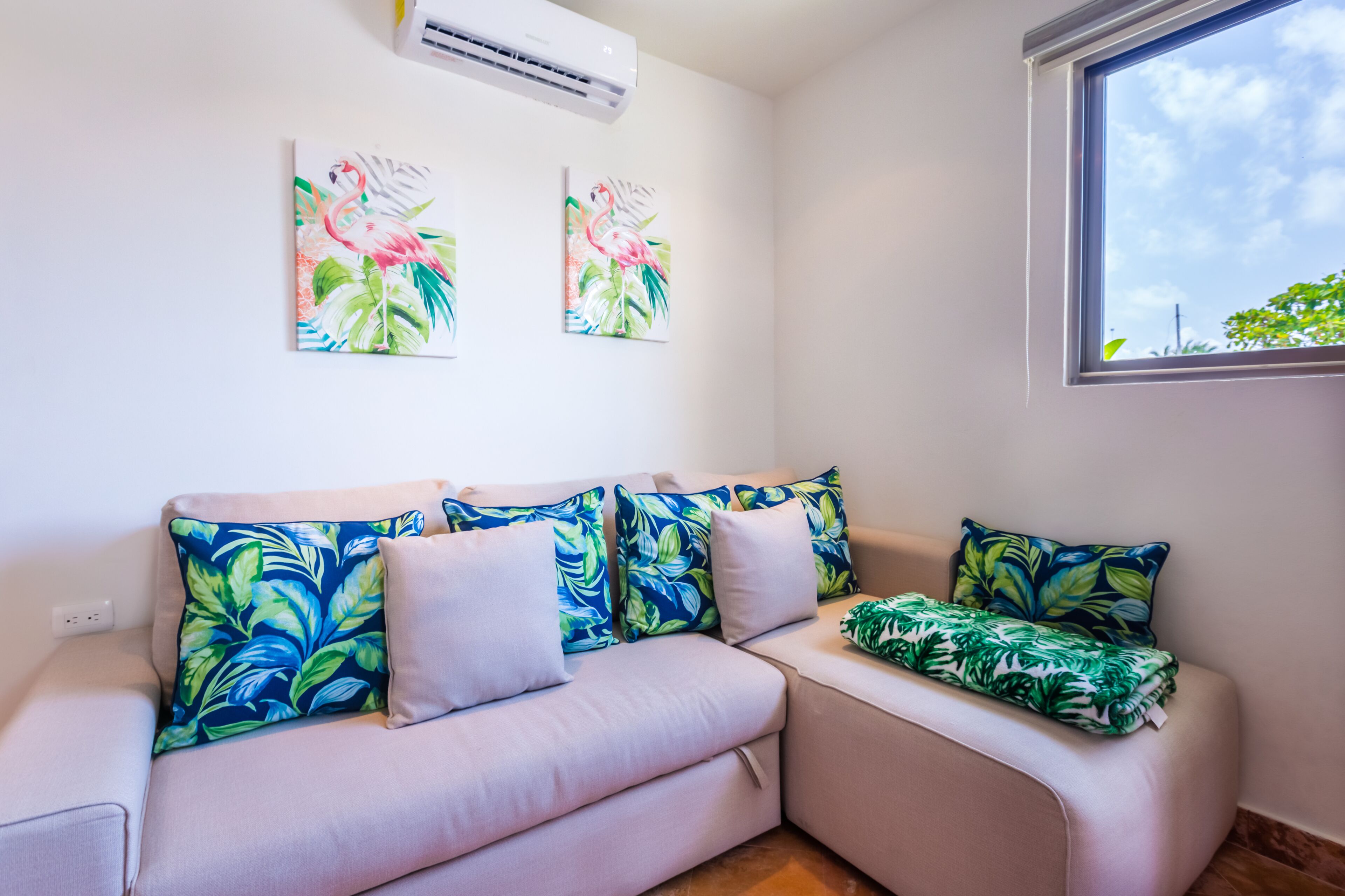 Un salon moderne avec un canapé d’angle beige, des coussins aux motifs tropicaux et deux tableaux représentant des flamants roses, accrochés au mur.