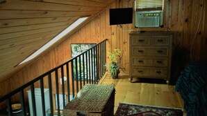 Un intérieur en bois avec un plafond en pente, un téléviseur fixé au mur, une commode, une plante et un tapis à motifs.