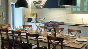 A dining area with a wooden table set for six, surrounded by chairs, with a kitchen in the background.