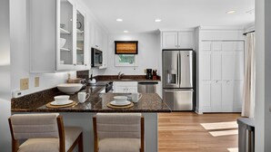 A modern kitchen with white cabinets, a granite countertop, and stainless steel appliances. There are two bar stools and a dining table. - Excellent Inlet Cove Cottage! Community Pool, Fishing Dock! (Johns Island)